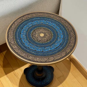 May include: A round side table featuring a black base and a circular top decorated with intricate, concentric mandala-style patterns in shades of metallic gold, deep black, and vibrant blue, supported by a central pedestal with a three-legged base.