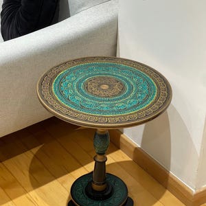 Puede incluir: Una mesa auxiliar redonda con un tablero circular que presenta intrincados patrones de estilo mandala en turquesa, dorado y negro. La mesa se apoya sobre una base de pedestal central con una plataforma circular a juego en tonos oscuros.