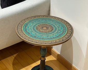 May include: A round side table featuring a circular top with intricate teal, gold, and black mandala-style patterns. The table stands on a central pedestal base with a matching circular platform, finished in coordinating dark tones and gold accents.