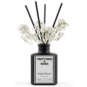 May include: A matte black glass reed diffuser bottle with a white label reading PRETTYMINI & MAGIC, Naked Sheets, Beach Linen + White Tea, Diffuser, 100g. Four black reeds are inserted into the bottle, surrounded by white baby's breath flowers.