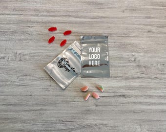 May include: Two silver zippered pouches with a white background. The pouch on the left has the text "Moonshine Designs" printed on it. The pouch on the right has the text "Your Logo Here" printed on it. There are red and pink candies scattered around the pouches.