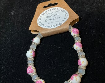 Pode incluir: Pulseira elástica branca com contas florais rosa e douradas e contas transparentes brilhantes. A pulseira tem uma etiqueta feita à mão que diz "Handmade Stretch Bracelet By Amanda".