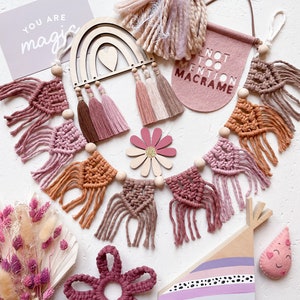 May include: A selection of handmade macrame and wooden decorations in pink, brown, and beige. Includes a rainbow, garland, flower, and a banner reading "NOT TOO MENTION MACRAME". A card says "YOU ARE magic".