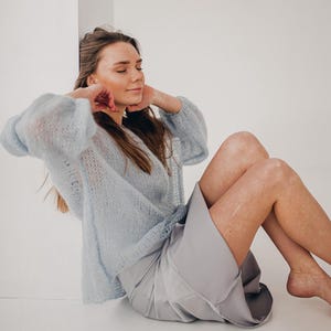 Könnte beinhalten: Hellblauer, durchsichtiger Strickpullover mit voluminösen Ärmeln und einem passenden hellgrauen Rock. Das Model sitzt mit geschlossenen Augen, Händen am Gesicht und ausgestreckten Beinen. Das Outfit ist weich und bequem.