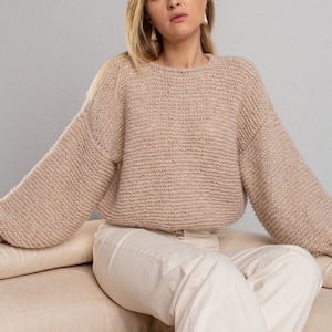 May include: A woman wearing a light brown knitted jumper with long sleeves and white trousers sits on a light brown sofa.