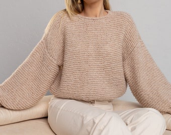 May include: A woman wearing a light brown knitted sweater with long sleeves and white pants sits on a light brown couch.