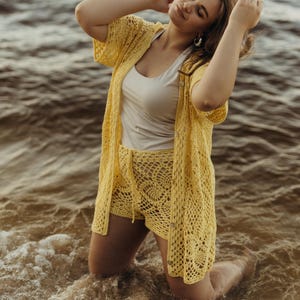May include: A yellow crochet cardigan, shorts, and a white tank top. The open-weave cardigan has short sleeves and button closures. The shorts have a drawstring waist. The outfit is worn in the ocean.