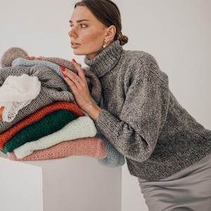 May include: A stack of folded sweaters in various colors, including gray, orange, green, and pink, rests on a white surface. A woman wearing a gray turtleneck sweater leans against the stack. The sweaters appear to be made of a soft, knitted material.