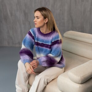 May include: A woman wearing a purple and blue striped sweater sits on a white couch. She is wearing white pants and brown boots.