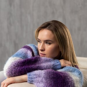 May include: A woman wearing a purple and blue striped sweater, resting her arms on a white couch. She is looking off to the side with a thoughtful expression.
