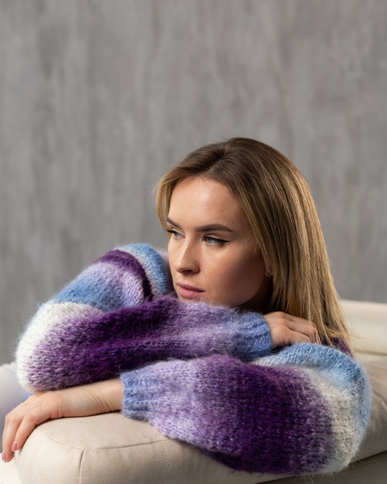May include: A woman wearing a purple and blue striped sweater, resting her arms on a white couch. She is looking off to the side with a thoughtful expression.
