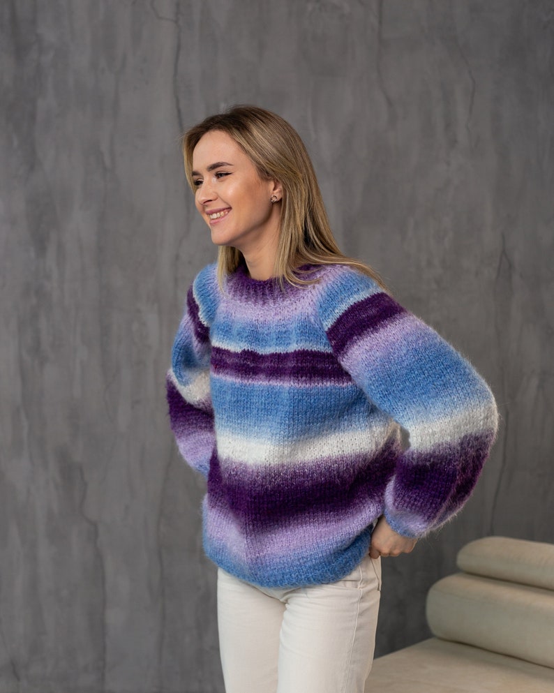 May include: A woman wearing a purple, blue, and white striped fuzzy sweater with long sleeves. She is smiling and looking to the right.