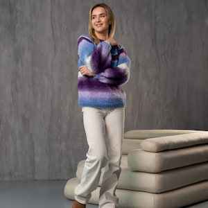 May include: A woman wearing a purple and blue striped fuzzy sweater with white pants and brown boots. She is standing in front of a white couch.