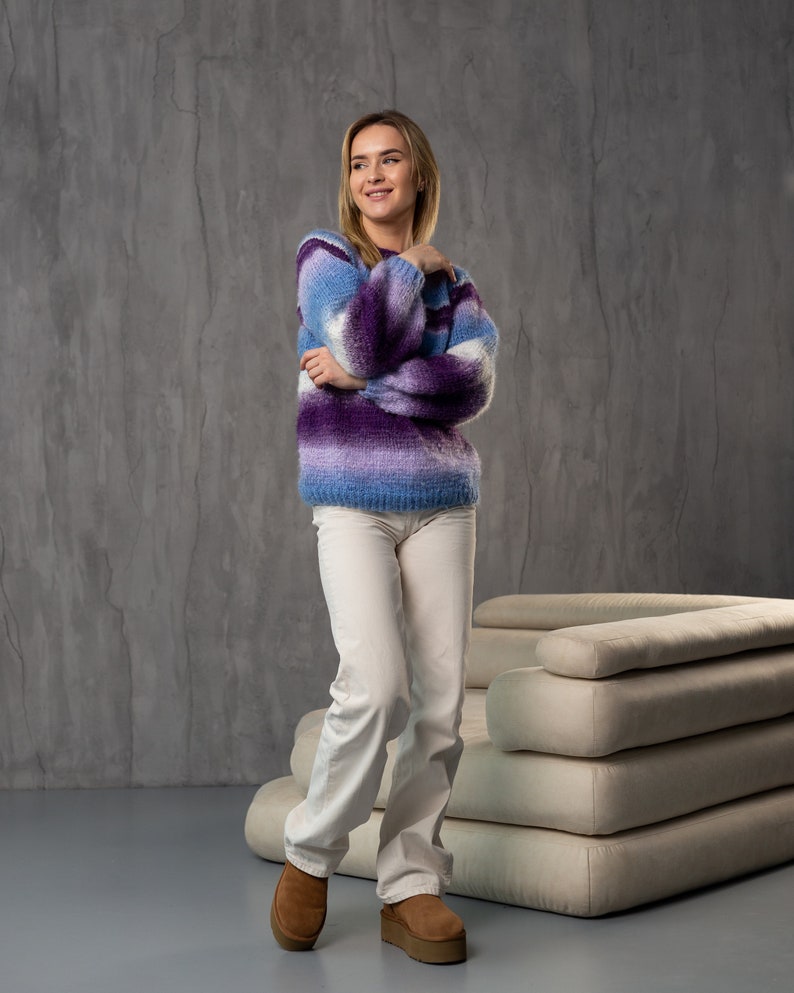May include: A woman wearing a purple and blue striped fuzzy sweater with white pants and brown boots. She is standing in front of a white couch.