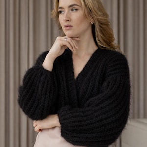 May include: A black, chunky-knit cardigan with a V-neck and long, voluminous sleeves. The sweater is worn over a light pink garment. The overall aesthetic is cozy and stylish, suitable for fall or winter wear.