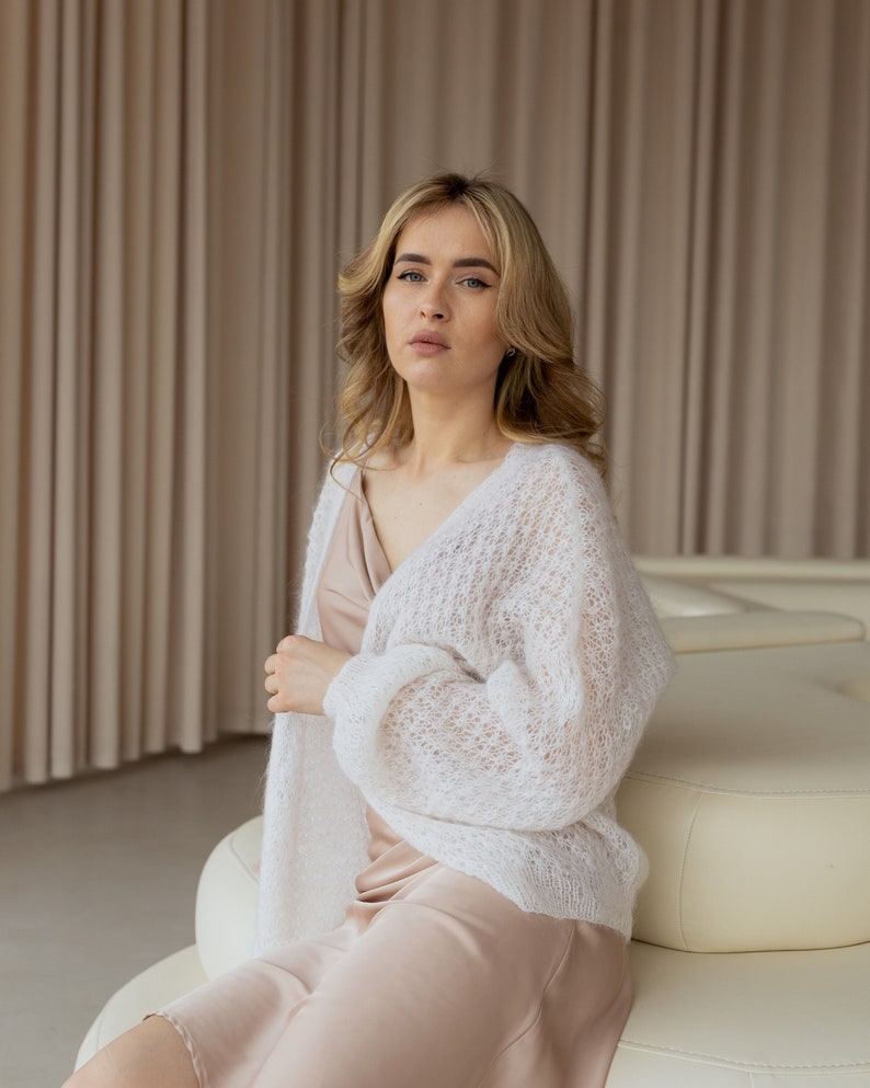 May include: A woman wearing a white knit cardigan over a beige slip dress. She is sitting on a white couch in a room with beige curtains.