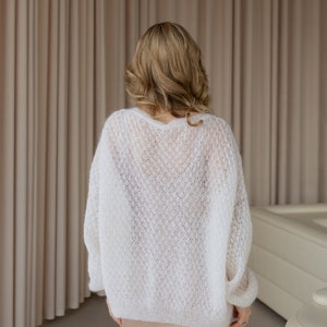 May include: A white, open-knit sweater with a loose fit. The sweater is made of a soft, delicate yarn and has a relaxed, casual style.