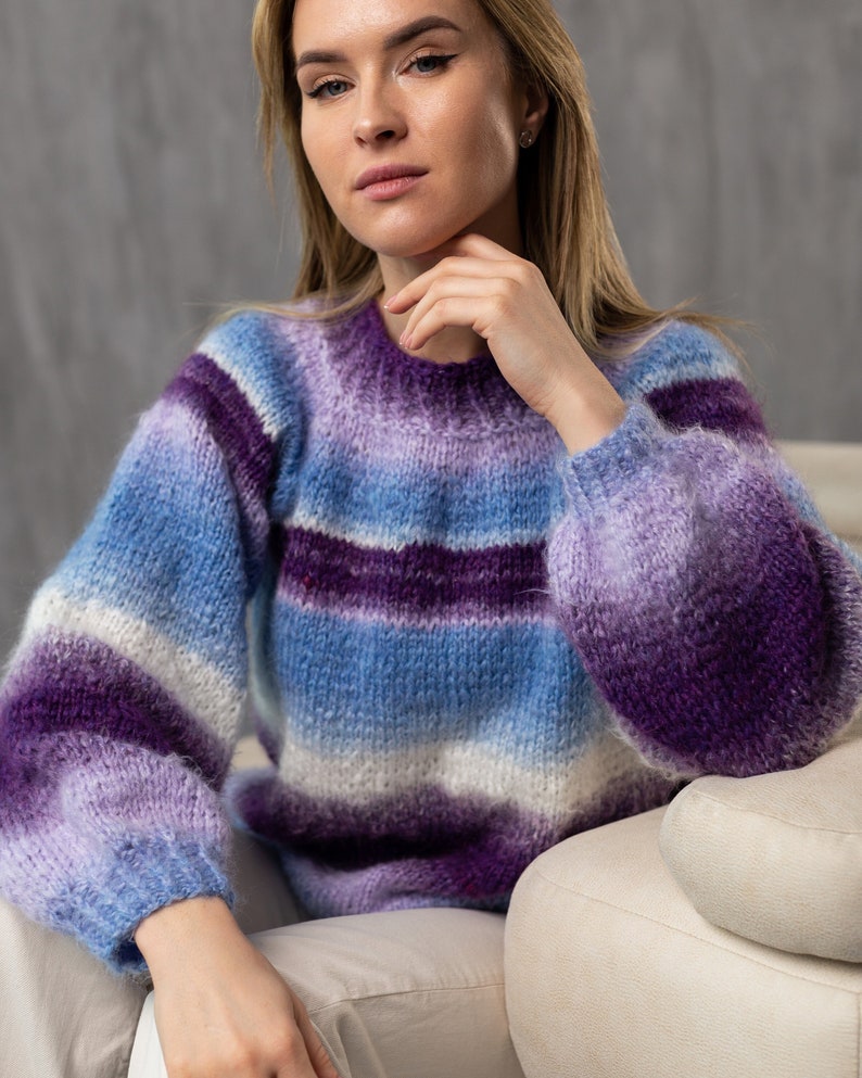 May include: A woman wearing a purple and blue striped sweater with a fuzzy texture. The sweater has a relaxed fit and is made of a soft, comfortable material.