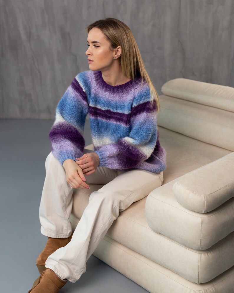 May include: A woman wearing a purple and blue striped sweater sits on a white couch. She is wearing white pants and brown boots.