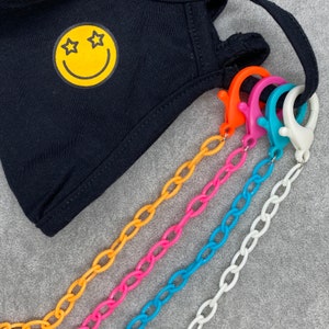 May include: Five colorful plastic chains with lobster clasps attached to a black face mask with a yellow smiley face with stars for eyes.