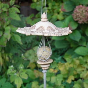 Könnte beinhalten: Ein Keramik-Vogelhäuschen mit einem weißen und braunen Design. Der Futterspender hat ein dekoratives Oberteil und einen hängenden Käfig für einen Fettball. Der Käfig ist aus Schnur und der Fettball ist im Inneren sichtbar.