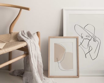 Könnte beinhalten: Minimalistische Innenszene mit einem Holzstuhl, einem Leinenüberwurf und gerahmten Kunstdrucken. Die Drucke zeigen eine Strichzeichnung einer Frau mit Hut und ein abstraktes Design in neutralen Tönen.