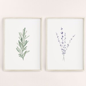 K&ouml;nnte beinhalten: Zwei gerahmte botanische Drucke. Einer zeigt einen Aquarellzweig gr&uuml;nen Rosmarins. Der andere Druck zeigt einen Aquarellstrau&szlig; lila Lavendel. Beide Drucke haben einen wei&szlig;en Hintergrund und sind in hellen Rahmen.