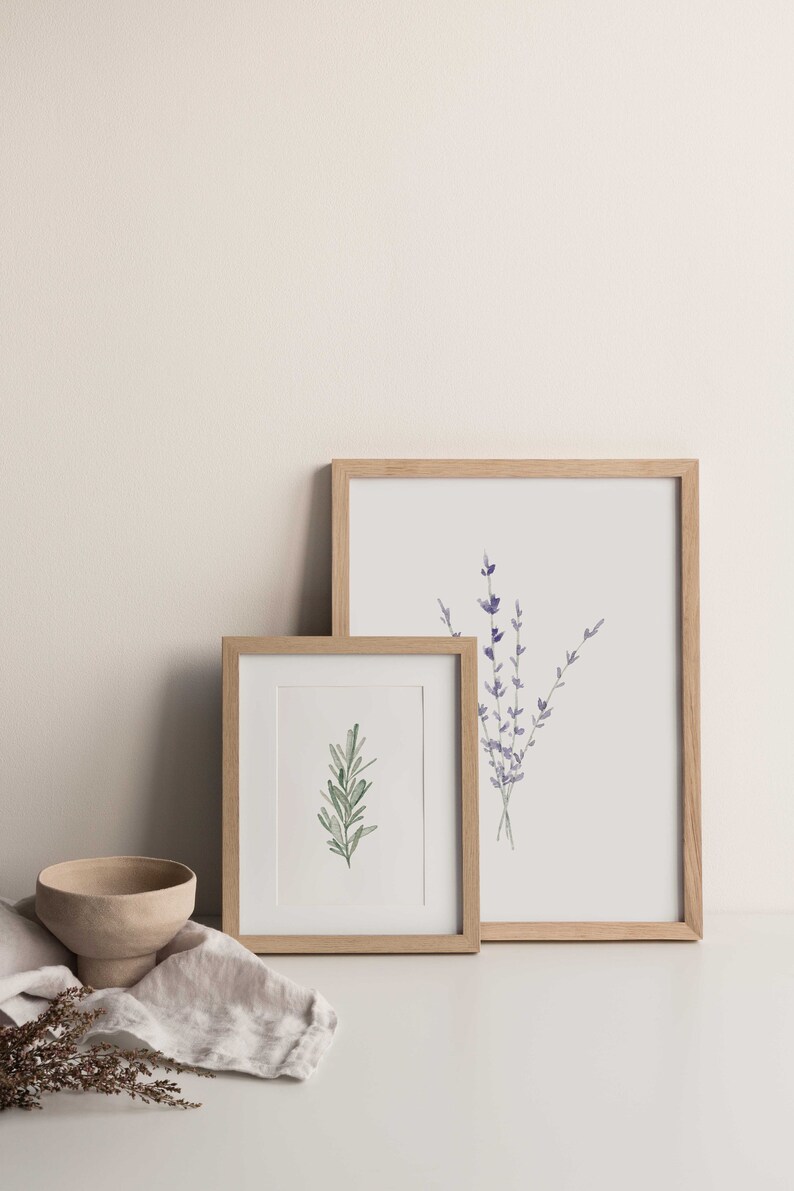 K&ouml;nnte beinhalten: Zwei gerahmte botanische Drucke mit hellen Holzrahmen. Ein Druck zeigt einen Rosmarinzweig, der andere einen Lavendelstrau&szlig;. Eine kleine, beige Schale und ein Leinentuch befinden sich im Vordergrund.