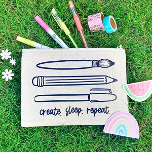 May include: A beige canvas pencil case with a zipper, featuring illustrations of a paintbrush, pencil, and pen, along with the text "create. sleep. repeat." The case is filled with colorful pens and washi tape, and is surrounded by decorative stickers.