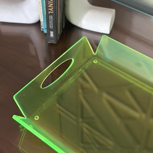 May include: A pair of white hand-shaped bookends holding a stack of books, including "Booze and Vinyl". A neon green rectangular tray with handle cutouts is in the foreground. The items are on a dark brown surface.