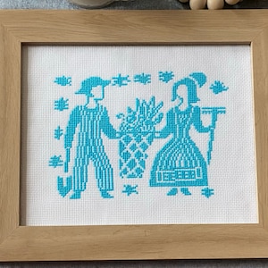May include: A framed cross-stitch embroidery depicting two figures in blue, one holding a bouquet of flowers and the other carrying a rake. The background is white with a simple design of stars.