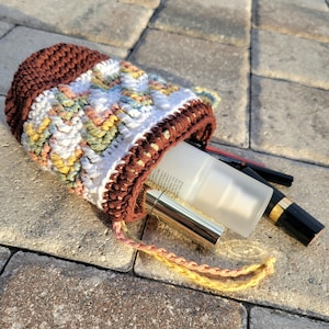 May include: A handmade crochet drawstring bag in brown, white, and pastel colors. The bag contains a tube of lotion, a lipstick, and other cosmetic items. The bag is sitting on a stone surface.