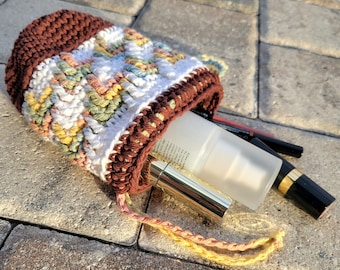 Puede incluir: Un bolso de ganchillo hecho a mano con cordón en colores marrón, blanco y pastel. El bolso contiene un tubo de loción, un lápiz labial y otros artículos cosméticos. El bolso está sobre una superficie de piedra.