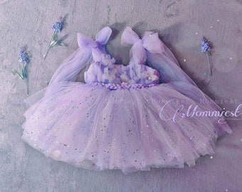 May include: A lavender tulle dress with a sparkly bodice and a bow on each shoulder. The dress has a braided waistband and is decorated with flowers. The dress is on a light gray background.