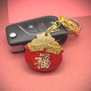 以下が含まれることがあります： 「福」の漢字が入った赤と金のキーホルダーが、黒い車のキーに付いています。キーホルダーは、赤いベースの上に金色のグリッターデザインが施されています。