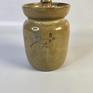 May include: A tan ceramic jar with a matching lid and a small knob handle. The jar features a speckled texture and a hand-painted floral design in blue and brown on the front. The vessel has a wide, flared rim and a rounded, cylindrical body.