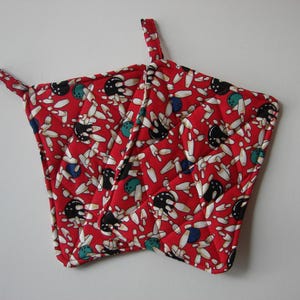 May include: Red quilted pot holder with a bowling theme. The fabric features a pattern of white bowling pins, black bowling balls, and teal and blue accents. The pot holder has a loop for hanging.