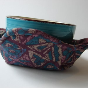 May include: A teal ceramic bowl with a brown rim sits inside a fabric bowl cover. The cover is made of a dark purple fabric with a teal and beige geometric pattern.