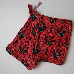 May include: Two red quilted pot holders with a black butterfly and insect pattern. The pot holders have a loop for hanging.