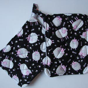 以下が含まれることがあります： 白い羊の模様とピンクのアクセントが付いた、黒いキルティングの鍋つかみが2つ。鍋つかみには吊り下げ用のループが付いています。