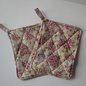 May include: Two quilted pot holders with floral patterns. The pot holders feature a cream-colored background with pink flowers and light blue ribbons. Each pot holder has a small loop for hanging.