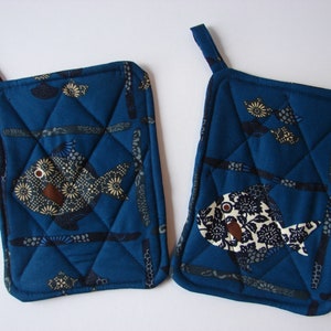 May include: Two square, quilted potholders in a deep blue fabric. Each features a fish applique with floral and geometric patterns in shades of cream, brown, and black. The potholders have a loop for hanging.