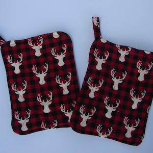 Puede incluir: Dos manoplas acolchadas con un estampado de cuadros rojos y negros. Cada manopla presenta un patrón repetido de cabezas de ciervo blancas. Las manoplas tienen un lazo para colgar.
