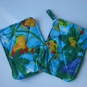 Könnte beinhalten: Zwei blaue, gesteppte Topflappen mit einem Frosch- und tropischen Blattdesign. Die Topflappen haben eine Schlaufe zum Aufhängen.