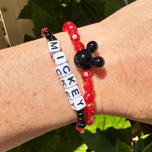 Könnte beinhalten: Zwei Armbänder, eines mit roten Perlen und einem schwarzen Mickey-Maus-Anhänger, das andere mit schwarzen Perlen und weißen Buchstabenperlen, die "MICKEY" buchstabieren.
