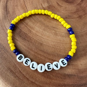 May include: A yellow and blue beaded bracelet with the word "BELIEVE" spelled out in white beads.