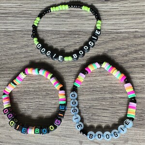 Puede incluir: Tres pulseras de cuentas con el texto "OOGIE BOOGIE". Una pulsera tiene cuentas negras y verde neón. Las otras dos pulseras tienen cuentas coloridas de arcoíris y cuentas negras. Las pulseras están sobre una superficie de madera.