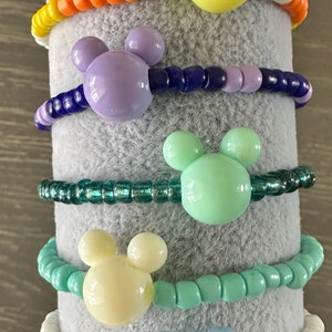 Pode incluir: Cinco pulseiras coloridas com pingentes de cabeça do Mickey Mouse. As pulseiras são feitas com pequenas contas de várias cores, incluindo laranja, azul, verde, verde claro e branco.