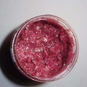 以下が含まれることがあります： キラキラ光る濃いピンク色の化粧品が入った透明なプラスチックジャー。製品は、虹色のグリッター粒子を含む、テクスチャード加工された粒状の外観をしています。ジャーは無地の白い背景に置かれています。
