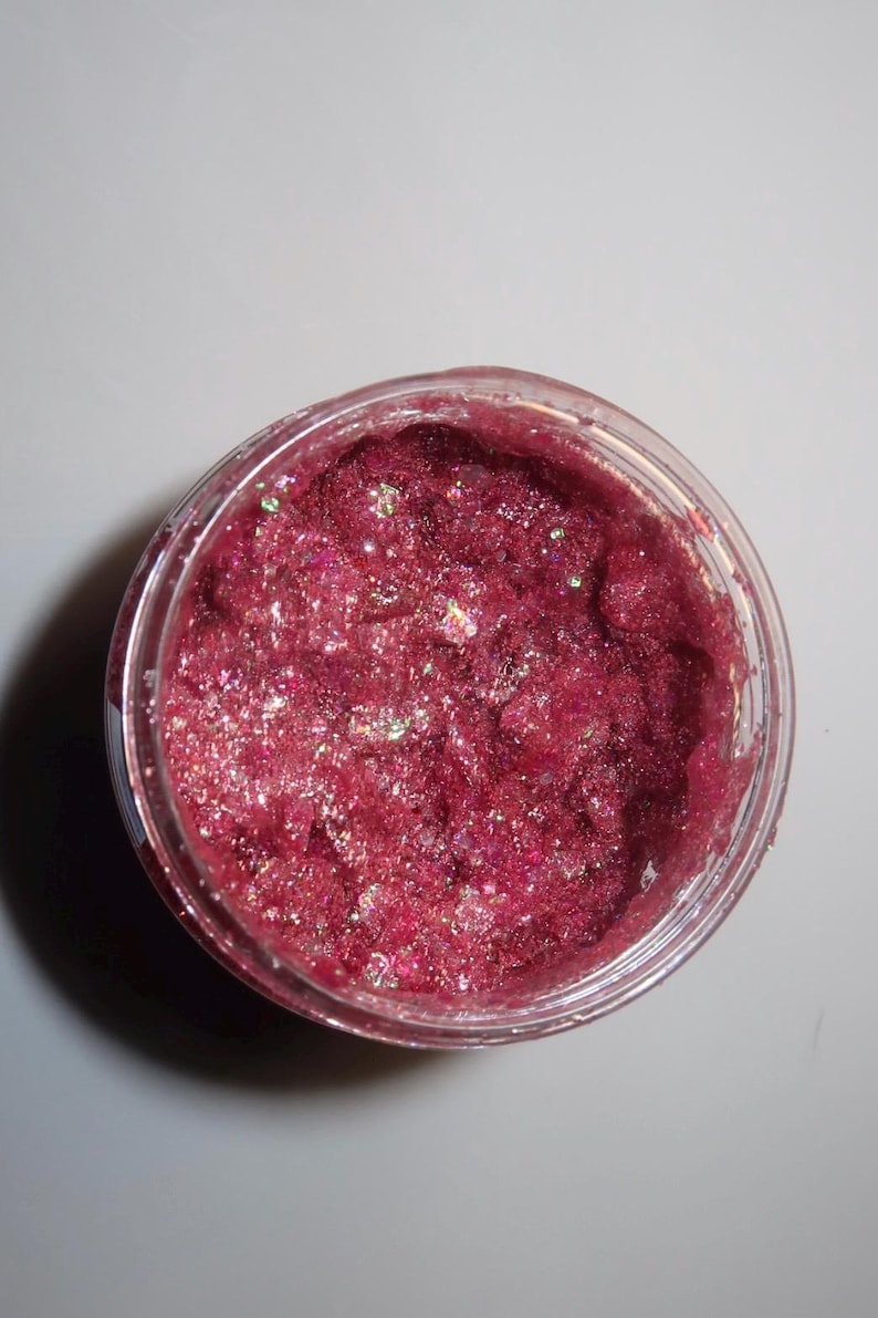 以下が含まれることがあります： キラキラ光る濃いピンク色の化粧品が入った透明なプラスチックジャー。製品は、虹色のグリッター粒子を含む、テクスチャード加工された粒状の外観をしています。ジャーは無地の白い背景に置かれています。